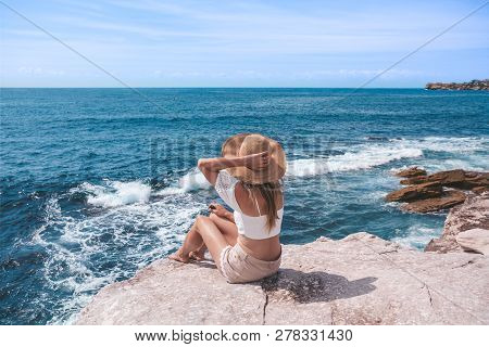 Woman Sitting By The Ocean Wearing Summer Crop Top And Shorts And A Sun Hat Watches The Waves Roll I