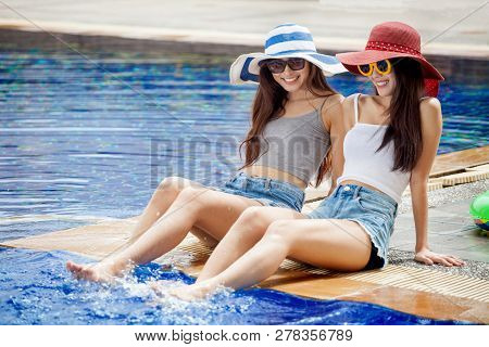 Two Beautiful Young Asian Women In Big Summer Hat And Sunglasses Sitting On The Edge Of The Swimming