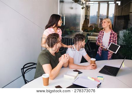 Serious Young Men Sit At Table And Look At Laptop Screen. Brunette And Blone Young Women Stand Behin