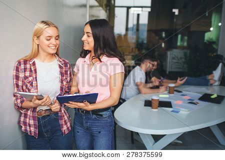 Cheerful And Positive Young Women Smile Together. They Stand In White Room. Models Hold Trye And Pla