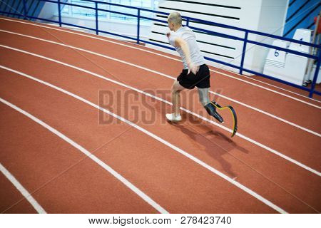 Young sportsman with prosthesis instead of right leg running down race track on stadium during competition