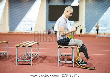 Paralympic competitor in activewear having break after workout at stadium and scrolling in smartphone for something to listen