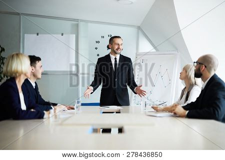 Confient business coach standing by table in front of employees and explaining his principles of successful work organization