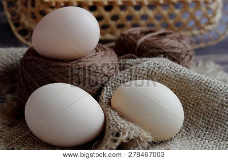 Three Eggs And Brown Threads Lie On A Beige Napkin On A Dark Background. Behind The Wicker Backgroun