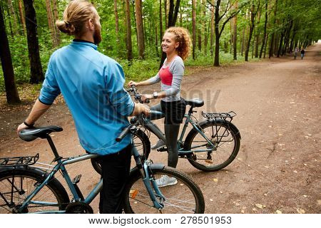 Happy young active couple with bicycles having talk while standing on forest road on summer deay