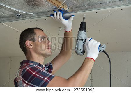 Young Man In Goggles Fixing Drywall Suspended Ceiling To Metal Frame Using Electrical Screwdriver On