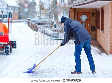 Man With Snow Shovel Cleans Sidewalks In Winter During Snowfall. Winter Time In Europe. Young Man In
