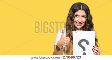 Young adult woman holding paper with question mark happy with big smile doing ok sign, thumb up with fingers, excellent sign