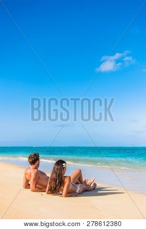Beach vacation couple tanning under the tropical sun enjoying summer holidays traveling aroung the world. Young people lying down in paradise. Vertical background with copy space on sky.