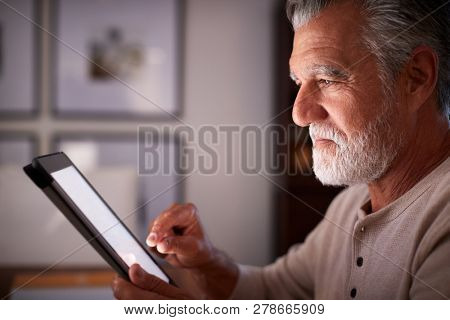 Senior Hispanic man sitting at a table using a tablet computer  in the evening, close up, side view