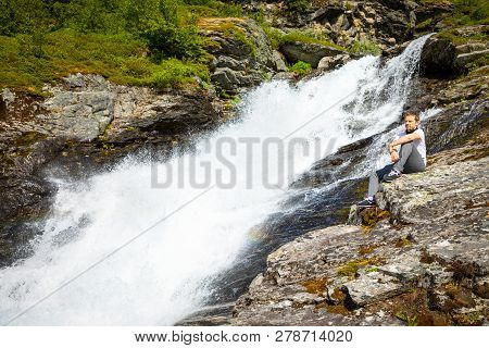 Young Man Next To Waterfall In The Geiranger Valley Near Dalsnibba Mountain In Norway