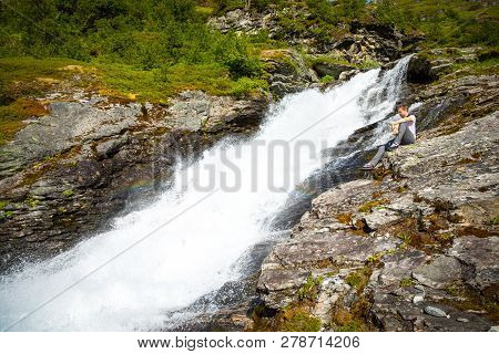 Young Man Next To Waterfall In The Geiranger Valley Near Dalsnibba Mountain In Norway