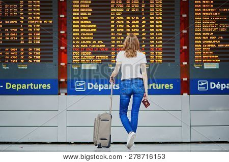 Young Woman In International Airport With Luggage And Passport Near Flight Information Display