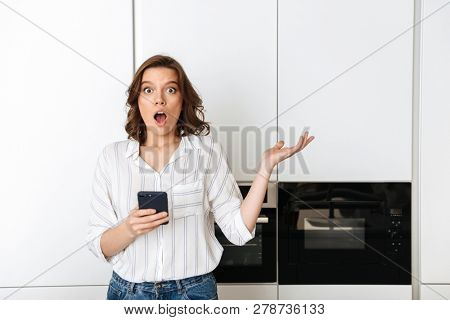 Shocked confused young businesswoman standing at the kitchen at home, holding mobile phone