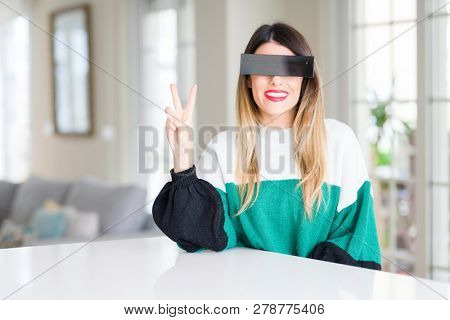 Young beautiful woman blindfold eyes with black censor band at home smiling with happy face winking at the camera doing victory sign. Number two.