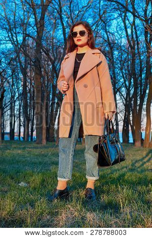 Beautiful Elegant And Stylish Young Brunette Woman, Wearing Sunglasses, Beige Coat And Jeans, Holdin