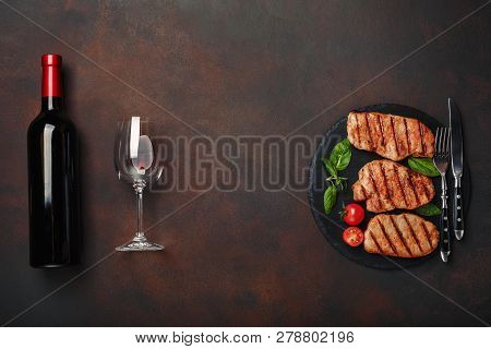 Grilled Pork Steaks On Stone With Bottle Of Wine, Wine Glass, Knife And Fork On Rusty Background