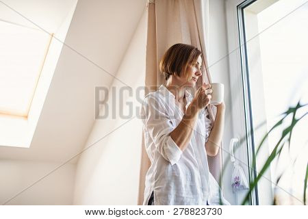 A Young Woman With Night Shirt Standing By The Window In The Morning, Holding Cup Of Coffee.