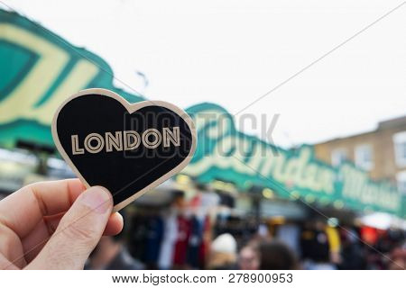 closeup of a caucasian man holding a black heart with the word london written in it, in front of the popular the camden market, in camden high street, in london, united kingdom