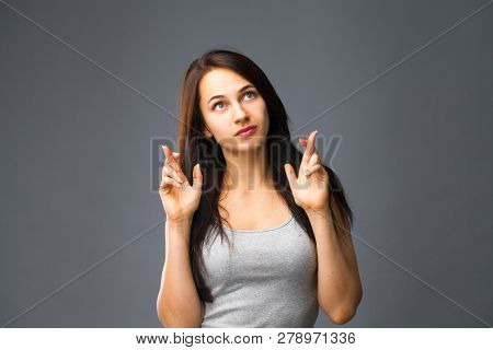 Body language and emotions concept. Glad dark haired beautiful satisfied girl beggs God to help her, keeps fingers crossed, wishes good luck, prays indoor isolated over gray studio background.