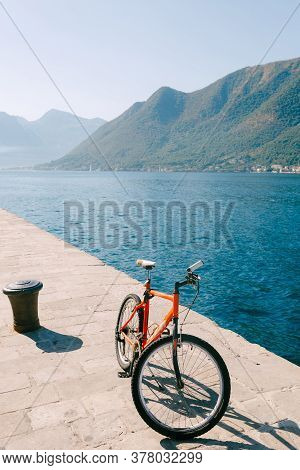 An Orange Bike On The Pier By The River Against The Green Mountains.