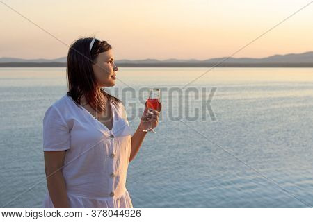 A Plump Girl Stands On The Shore Of A Lake With A Glass Of Wine At Sunset
