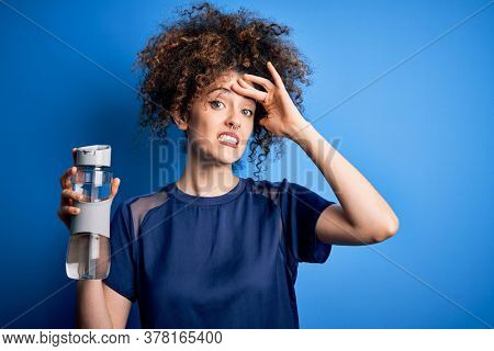 Young beautiful sporty woman with curly hair and piercing doing sport holding bottle of water stressed with hand on head, shocked with shame and surprise face, angry and frustrated. Fear and upse