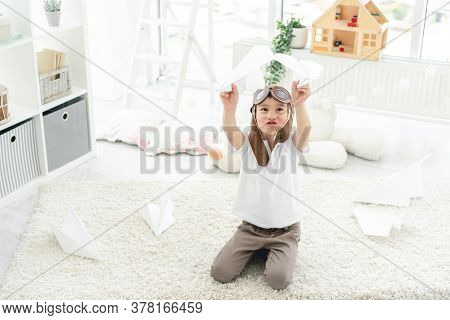 Funny little boy in pilot hat playing with paper planes in light kids room