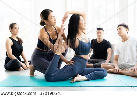 Asian people learning Yoga class in fitness club. Instructor coaching and adjust correct pose on king pigeon pose to student at front while others looking. Yoga Practice Work out lifestyle concept.