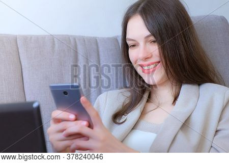 Woman Working Typing On Laptop And Reading Message On Smartphone Sitting On Couch Smiling, Distant W