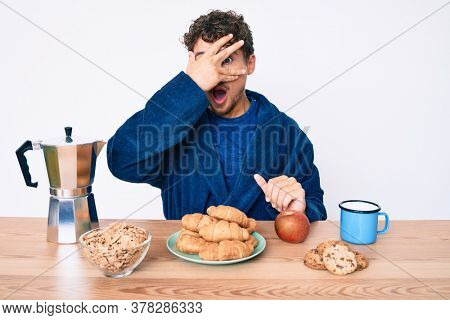 Young caucasian man with curly hair sitting on the table having breakfast peeking in shock covering face and eyes with hand, looking through fingers afraid 