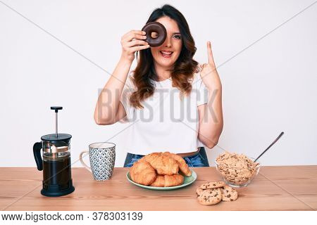 Young beautiful brunette woman eating breakfast holding cholate donut smiling with an idea or question pointing finger with happy face, number one 