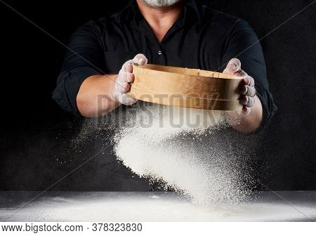 Chef A Man In A Black Uniform Holds A Round Wooden Sieve In His Hands And Sifts White Wheat Flour On
