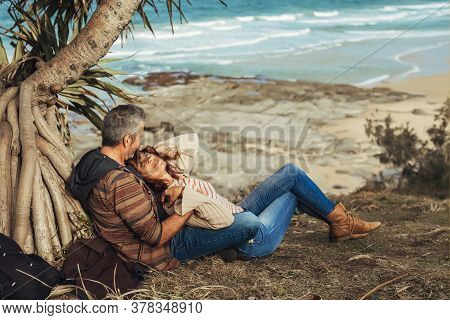 Happy Middle-aged Couple In Love Sitting Under The Tree With Ocen View, Hug, Smile And Look At Each 