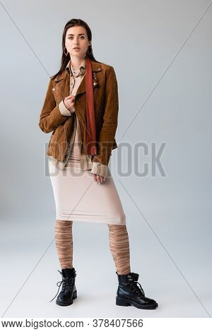 Full Length View Of Attractive Girl In Trendy Autumn Outfit Touching Suede Jacket On Grey