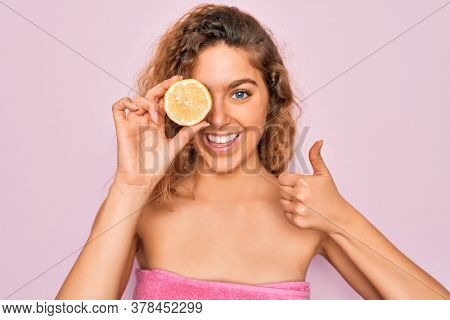 Beautiful woman with blue eyes wearing towel shower after bath holding lemon fruit over eye happy with big smile doing ok sign, thumb up with fingers, excellent sign