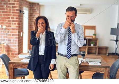 Two middle age business workers standing working together in a meeting at the office smelling something stinky and disgusting, intolerable smell, holding breath with fingers on nose. Bad smells.