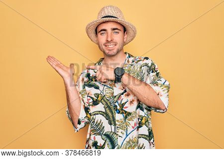 Young handsome man with blue eyes on vacation wearing summer florar shirt and hat amazed and smiling to the camera while presenting with hand and pointing with finger.