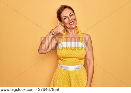 Middle age senior pin up woman wearing 50s style retro dress over yellow background smiling doing phone gesture with hand and fingers like talking on the telephone. Communicating concepts.