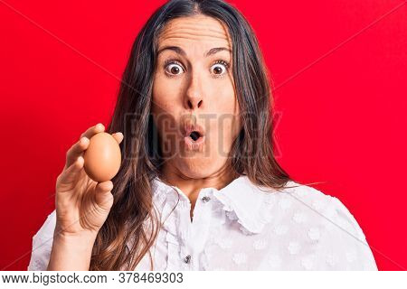 Beautiful brunette businesswoman holding bunch of dollars banknotes over red background scared and amazed with open mouth for surprise, disbelief face