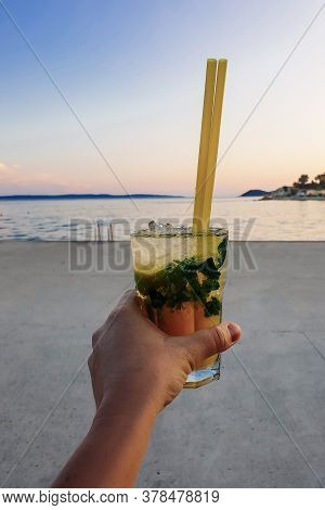 Closeup Of Womans Hand Holding Chilled Mohito Coctail With Drinking Straw And Ice Cubes.