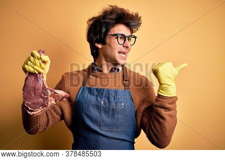 Young handsome butcher man holding meet steak standing over isolated yellow background smiling with happy face looking and pointing to the side with thumb up.