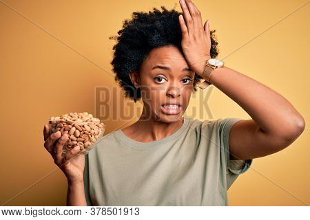 Young African American afro woman with curly hair holding bowl with salty peanuts stressed with hand on head, shocked with shame and surprise face, angry and frustrated. Fear and upset for mistake.