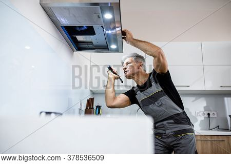 Aged Repairman In Uniform Working, Examining Broken Kitchen Extractor Fan, Using Flashlight. Repair 