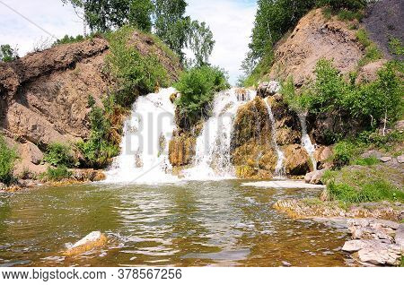 A Small Picturesque Waterfall Flowing Down Between The Mountains, Forming A Reservoir Surrounded By 