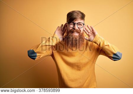 Handsome Irish redhead man with beard wearing glasses over yellow isolated background Trying to hear both hands on ear gesture, curious for gossip. Hearing problem, deaf
