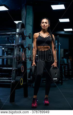 Full Length Portrait Of Fitnesswoman With Braids Posing Near Stand With Sports Equipment And Holding