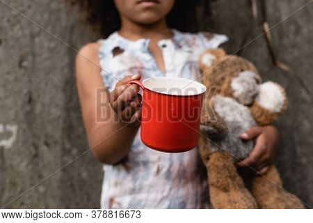 Cropped View Of Poor African American Kid Holding Metal Cup And Teddy Bear While Begging Alms On Urb