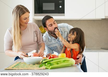 Family Cooking And Eating At Home During Pandemic. Girl Giving Slice Of Veg To Dad For Taste While M
