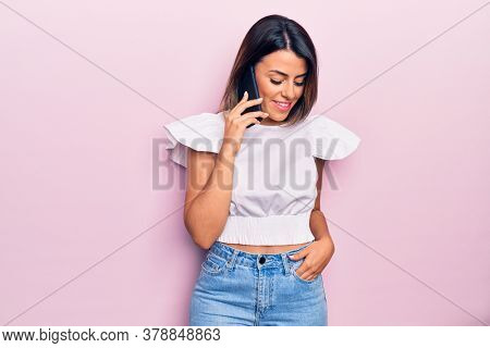 Young beautiful brunette woman smiling happy and confident. Having conversation talking on the smartphone standing with smile on face over isolated pink background